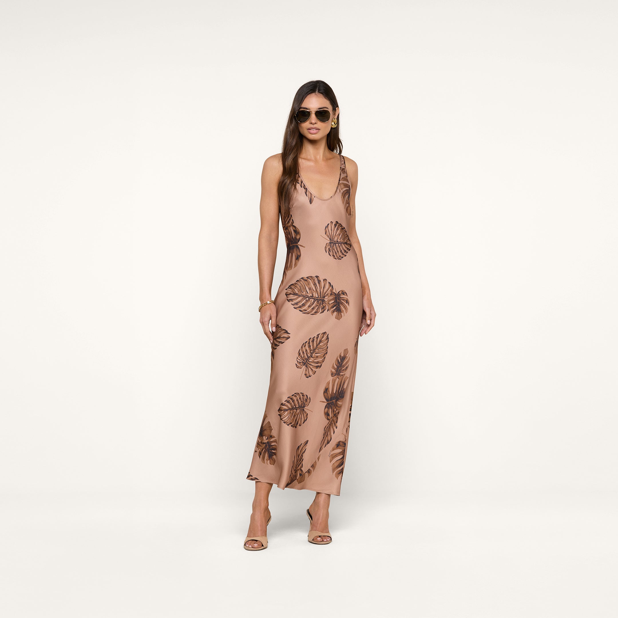 Woman wearing sunglasses and a sleeveless, ankle-length beige Akiya Satin Maxi Dress with a brown palm leaf print, standing against a plain white background. She has long hair and is wearing heeled sandals.