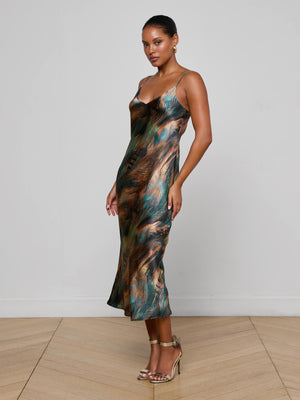 A woman poses against a white wall, standing on a light wood floor in the L'AGENCE Seridie Silk Slip Dress—bias-cut with a multicolored swirl pattern—and completes her look with gold strappy heels and hoop earrings.