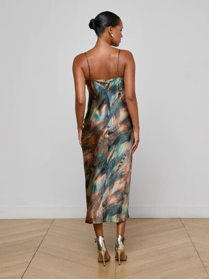 A woman, hair in a neat bun, faces away from the camera wearing the L'AGENCE Seridie Silk Slip Dress with a teal feather print and metallic heels, standing on a light wooden floor against a plain wall.
