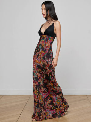 A woman with long, straight dark hair stands on a wooden floor wearing the L'AGENCE Susanna Silk Lace Gown—a long, flowing dress featuring a black bodice and colorful paisley print skirt. She faces to the side.