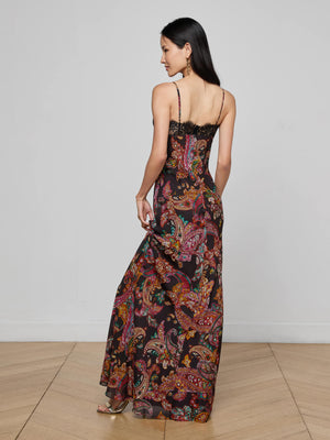 A woman with long dark hair wears the Susanna Silk Lace Gown by L’AGENCE, a floor-length dress with a colorful paisley print. She stands on a wooden floor, facing away to her left and holding part of the gown.