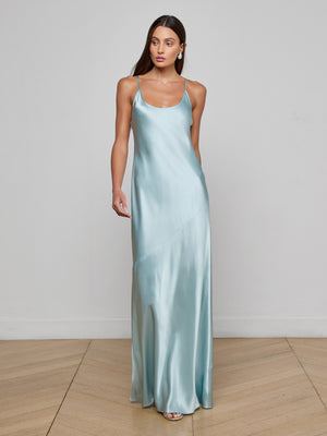 A woman with long brown hair stands on a wooden floor against a plain white wall, wearing the Charmaine Silk Gown—a light blue satin slip dress with thin straps. She looks slightly to the side with one hand by her side.