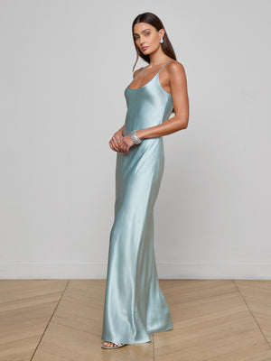 A woman stands on a wooden floor wearing the Charmaine Silk Gown—long, shiny, and light blue with thin straps. She accessorizes with silver jewelry and heels, posing by a plain wall—a perfect choice for a wedding guest dress.