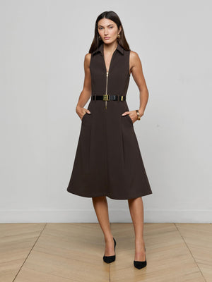 A woman stands against a light wall, wearing the sleeveless Kylo Shirt Dress by L’AGENCE in dark brown suiting fabric with a front zipper, black belt with gold buckle, black heels, and gold hoop earrings. She casually rests her hands in the dress’s pockets.
