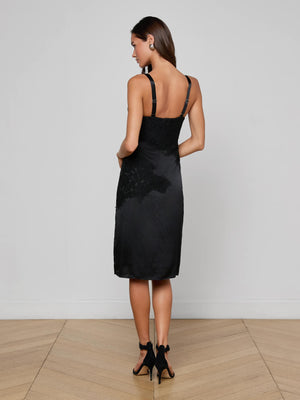 A woman stands on a wooden floor facing away, wearing the L'AGENCE Fenna Silk Lace Dress—a black silk charmeuse bustier dress with floral lace trim—paired with black high-heeled sandals against a plain white wall.