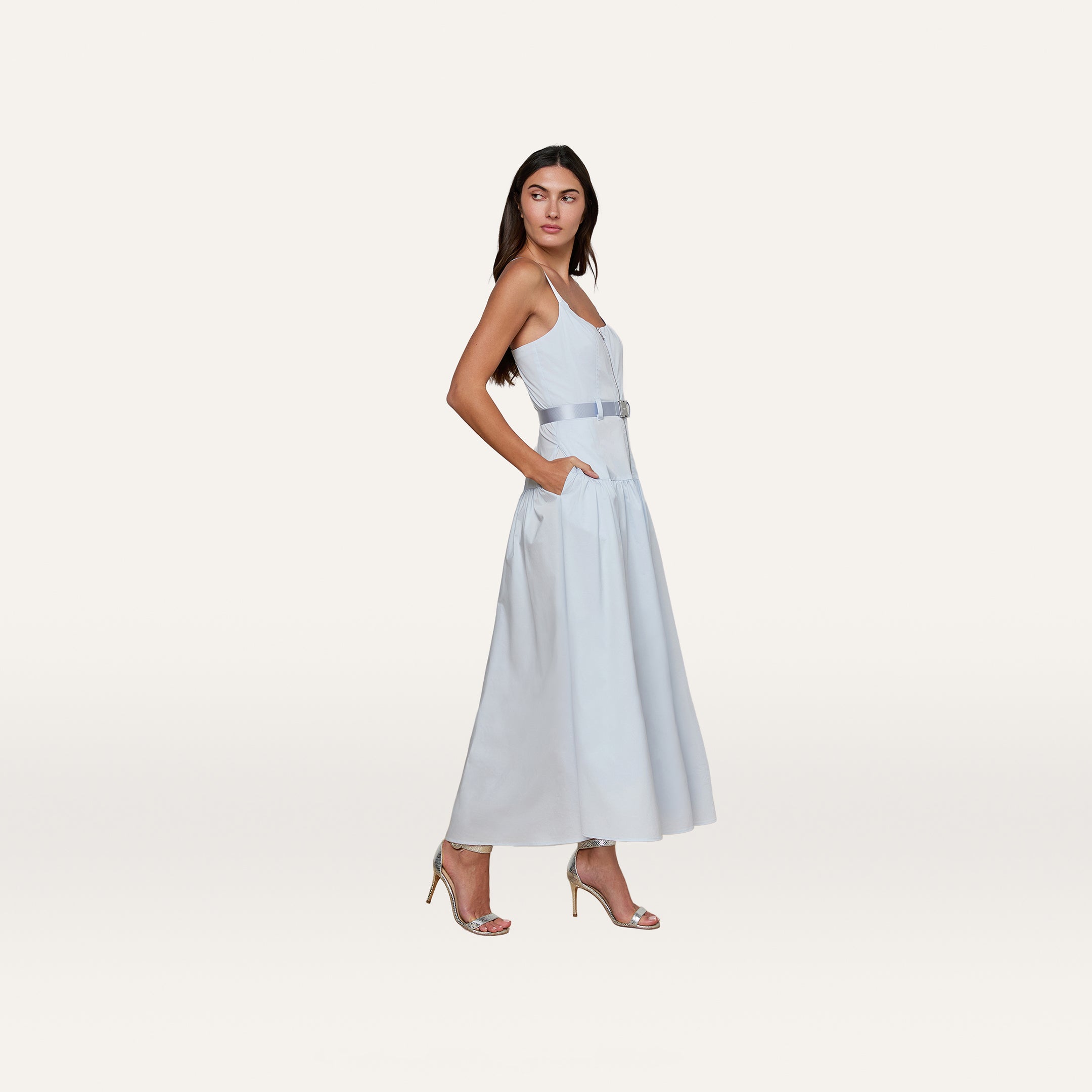 A woman stands sideways against a plain white background wearing the Rima Maxi Dress, a pale blue stretch poplin maxi style featuring a belt and pockets, paired with silver high-heeled sandals.