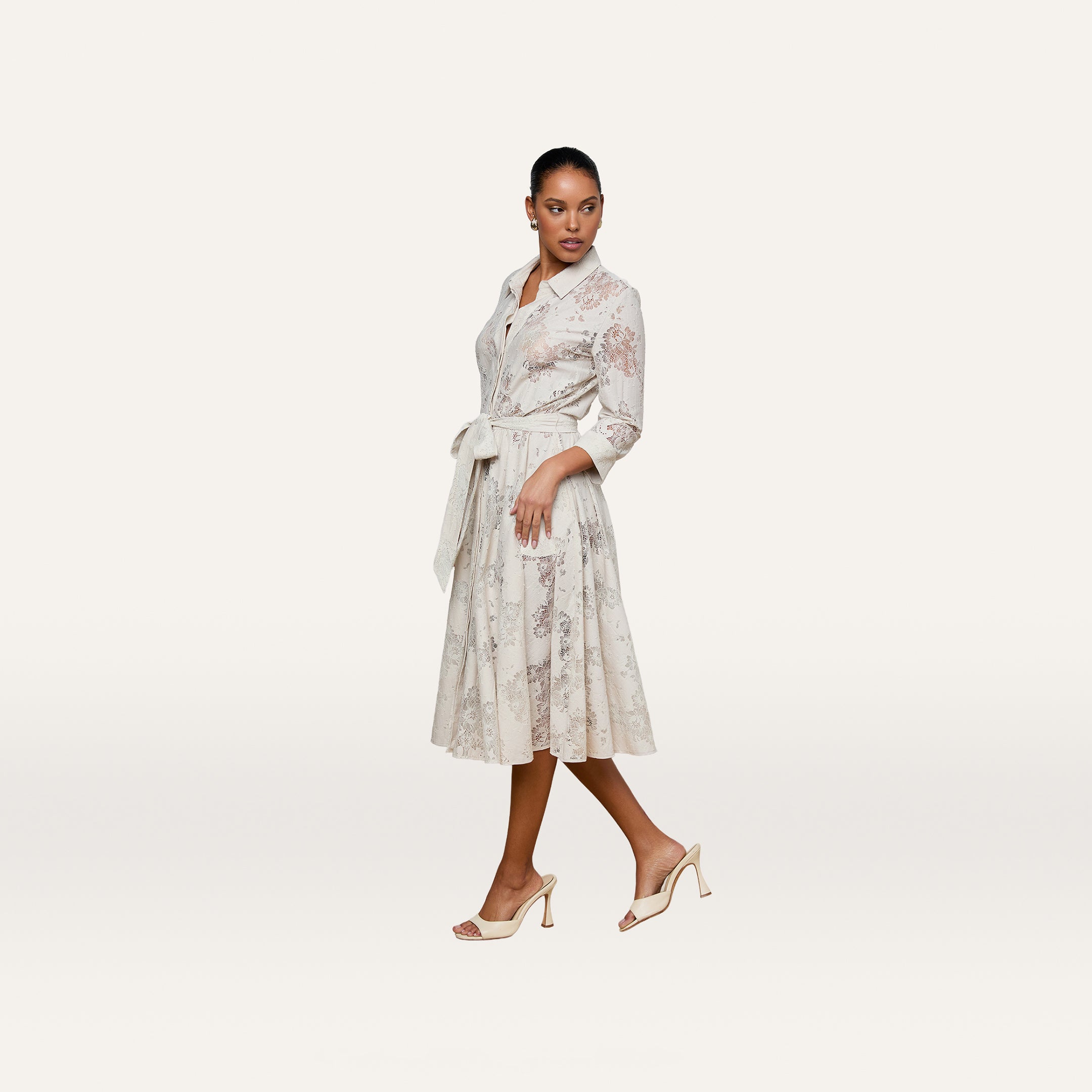 A woman is wearing the Ralphie Lace Midi Dress, featuring a classic silhouette with long sleeves, a belted waist, and a delicate floral pattern. She completes the look with light heels and stands against a plain white background.