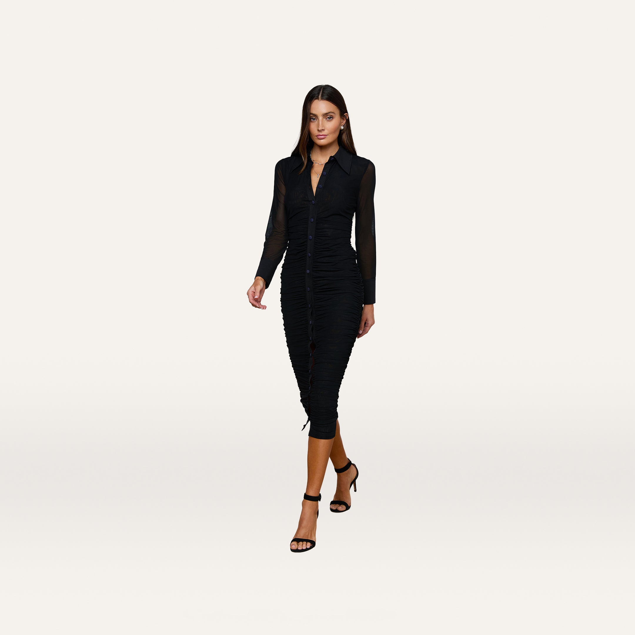 A woman wearing the Adriana Ruched Midi Dress—a long-sleeve, fitted black stretch mesh dress—paired with black heels, walking forward against a plain white background.