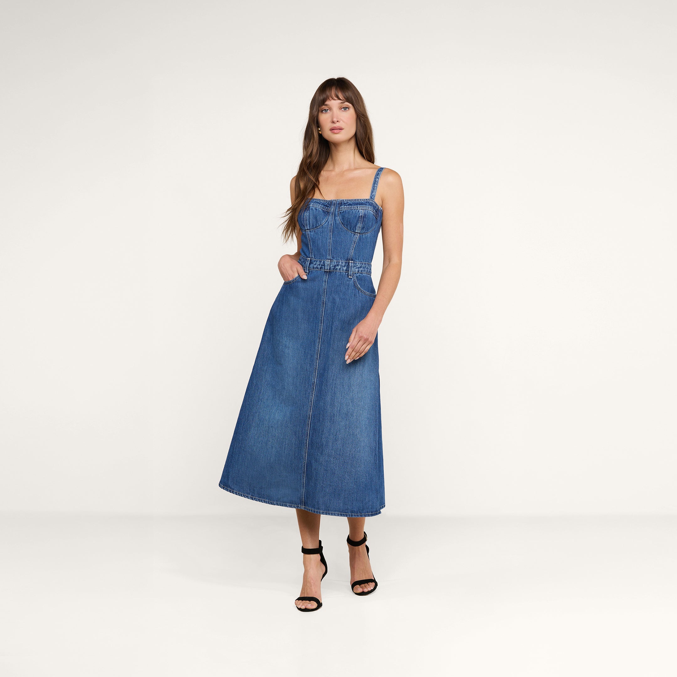 Sobre un fondo beige, una mujer de pelo largo y castaño luce el vestido «Jovie Bustier» —un vestido sin mangas de denim azul con corpiño tipo corsé y falda en línea A— combinado con unas sandalias negras de tacón alto, con una mano metida de forma desenfadada en el bolsillo.