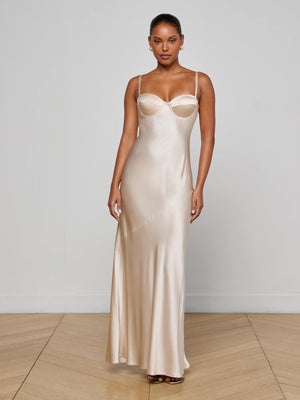 A woman stands indoors on a wooden floor, wearing the Sayla Silk Gown by L'AGENCE—a long champagne gown with thin straps and a fitted bodice. She exudes vintage glamour, facing the camera with arms relaxed by her sides against a plain light background.