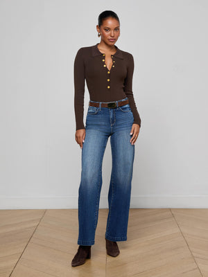A woman stands indoors against a plain light wall, wearing the Elisabetta Knit Bodysuit in chocolate brown with gold buttons, styled with blue jeans, a black belt, and brown heeled boots. She looks at the camera with a slight smile.