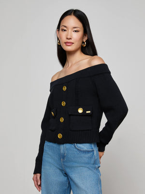 A woman with long dark hair wears the L'AGENCE Teri Off-The-Shoulder Top, a black cropped cardigan with gold buttons, paired with light blue jeans against a plain light gray background.