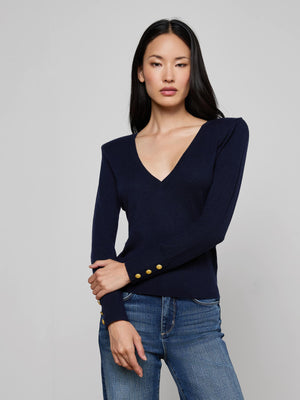 A woman with long black hair wears the Kolie Chain Top, a navy V-neck knit sweater with gold sleeve buttons, styled with blue jeans, standing against a light gray background.