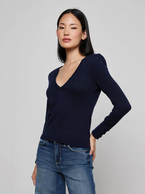 A woman with long dark hair, wearing the Kolie Chain Top—a refined navy knit sweater with a deep V-neckline—and blue jeans, stands against a plain light gray background. She has a neutral expression and rests one hand on her hip.