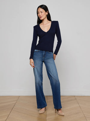 A woman with long dark hair poses indoors against a light wall, wearing the Kolie Chain Top in navy blue with a deep V-neckline, paired with blue jeans and beige heels; one arm rests by her side while the other is slightly bent.
