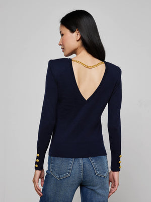 A woman with long dark hair models the Kolie Chain Top, a refined navy knit sweater with a deep V-back, high-shine chain detail, and gold button-accented sleeves. She completes the look with classic blue jeans.