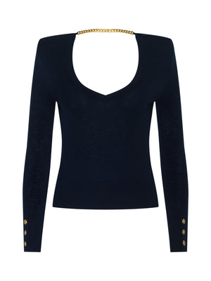 The Kolie Chain Top is a fitted black long-sleeve with structured shoulders, gold buttoned cuffs, a deep scoop neck, and a high-shine chain detail connecting the shoulders at the back.