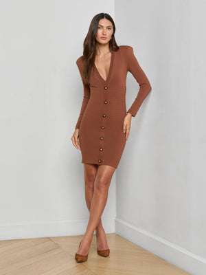 A woman stands indoors against a light gray wall, wearing the L'AGENCE Taru Button-Front Mini Dress with long sleeves, a deep V-neckline, and gold buttons. She has long, straight hair and pairs the look with brown heels.