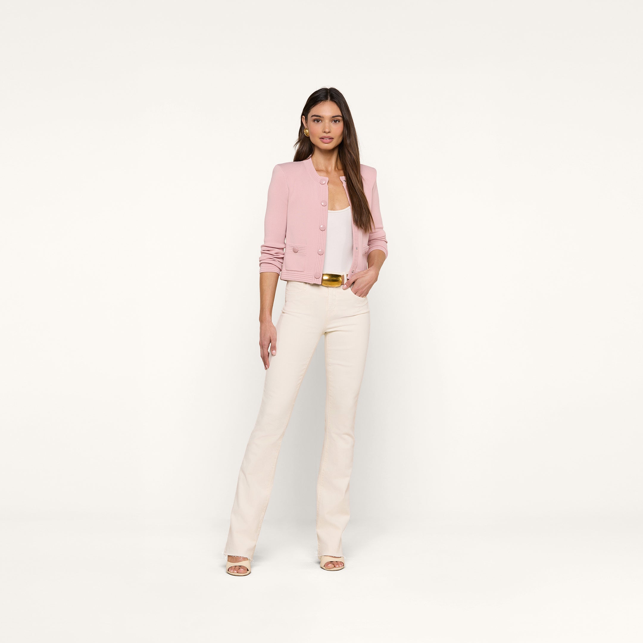 A woman stands against a plain white background, wearing the Patsy Cropped Cardigan in light pink over a white top, paired with cream-colored pants and open-toe heels. She has long brown hair and a neutral expression, with her hands by her sides. The cardigan features a subtle pointelle knit texture.