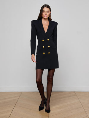 A woman with long brown hair and a neutral expression stands indoors against a plain light wall, wearing the L'AGENCE Meyumi Knit Blazer Dress, black sheer tights, and black heels.