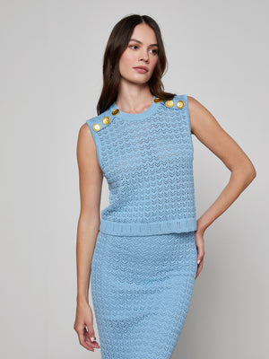 A woman with long brown hair and a neutral expression wears the Max Pointelle Knit Top in light blue, featuring gold shoulder buttons, along with a matching knit skirt, standing against a plain light gray background.