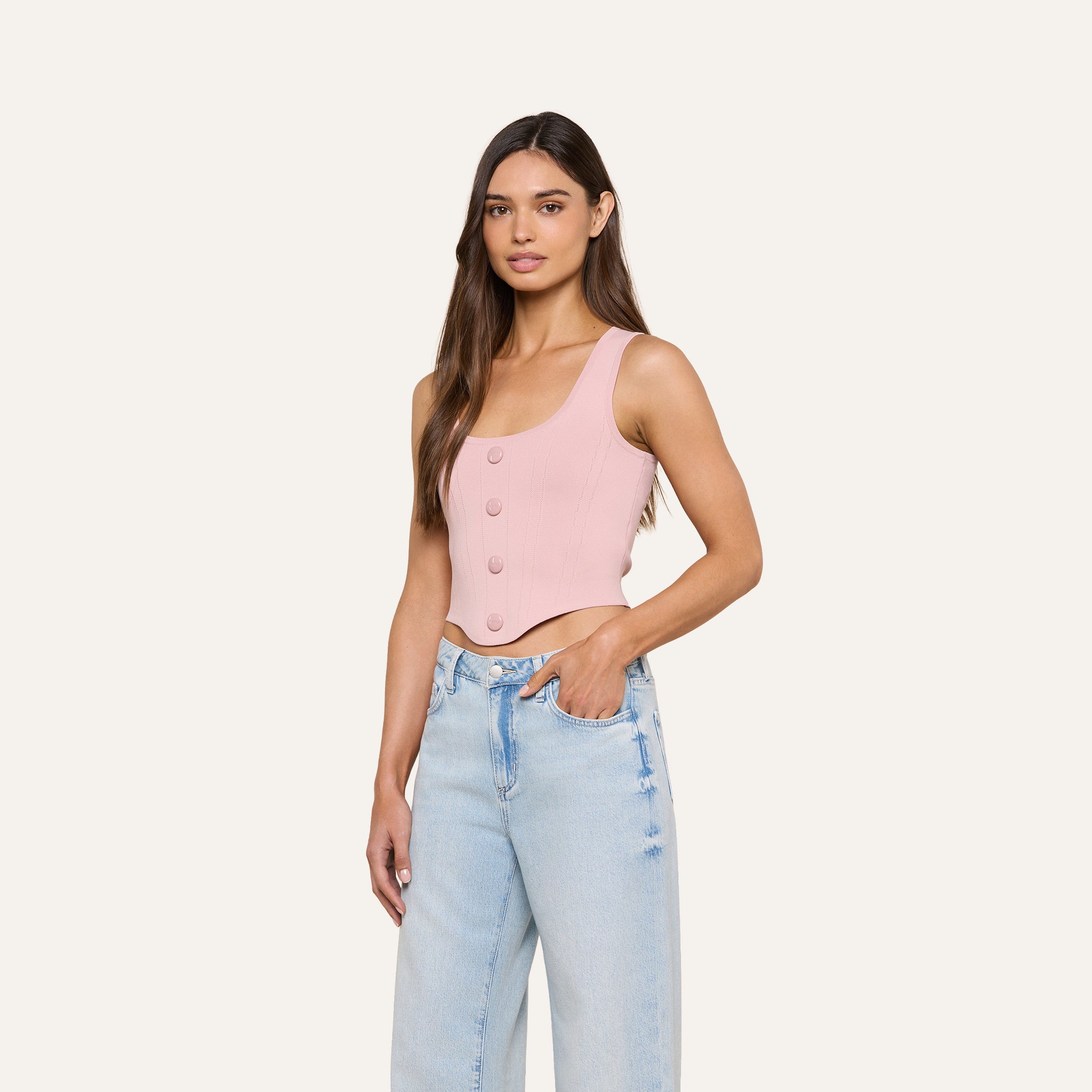 A woman wearing the Dove Knit Corset Top in light pink—a sleeveless, buttoned crop top with a curved hemline—paired with light blue high-waisted jeans, stands with one hand in her pocket against a plain off-white background.