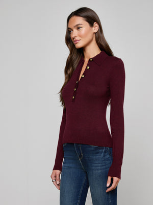 A woman models the Sterling Sweater—a fitted, long sleeve knit burgundy top with gold buttons—paired with blue jeans, standing against a plain light gray background and looking slightly to the side.