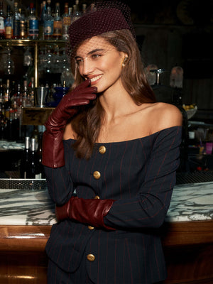 A woman beams with vintage style at a marble bar, wearing the Anstine Pillbox Hat with a birdcage veil, a pinstriped off-shoulder suit adorned with gold buttons, and red gloves. Bottles line the counter behind her.