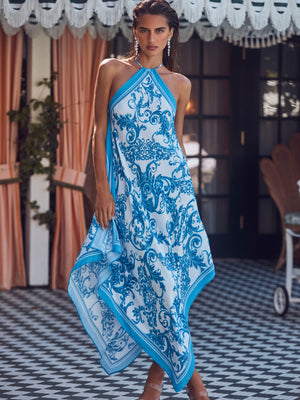 A woman stands on a checkered floor in the Elise Cover-Up Dress, a flowing blue baroque print halter midi dress with an ornate pattern. She is posed before glass doors framed by striped curtains and decorative trim above.