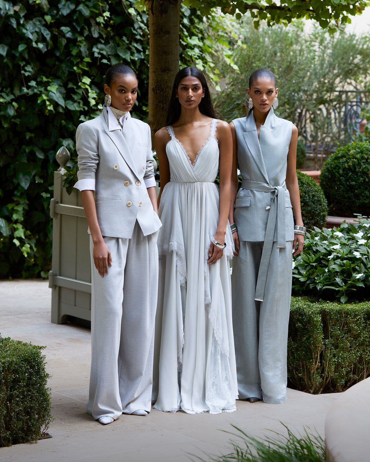 Three women pose outdoors in elegant, light-colored outfits: two in tailored suits with wide-leg pants, and one in a flowing lace-accented gown, surrounded by lush greenery and manicured bushes.