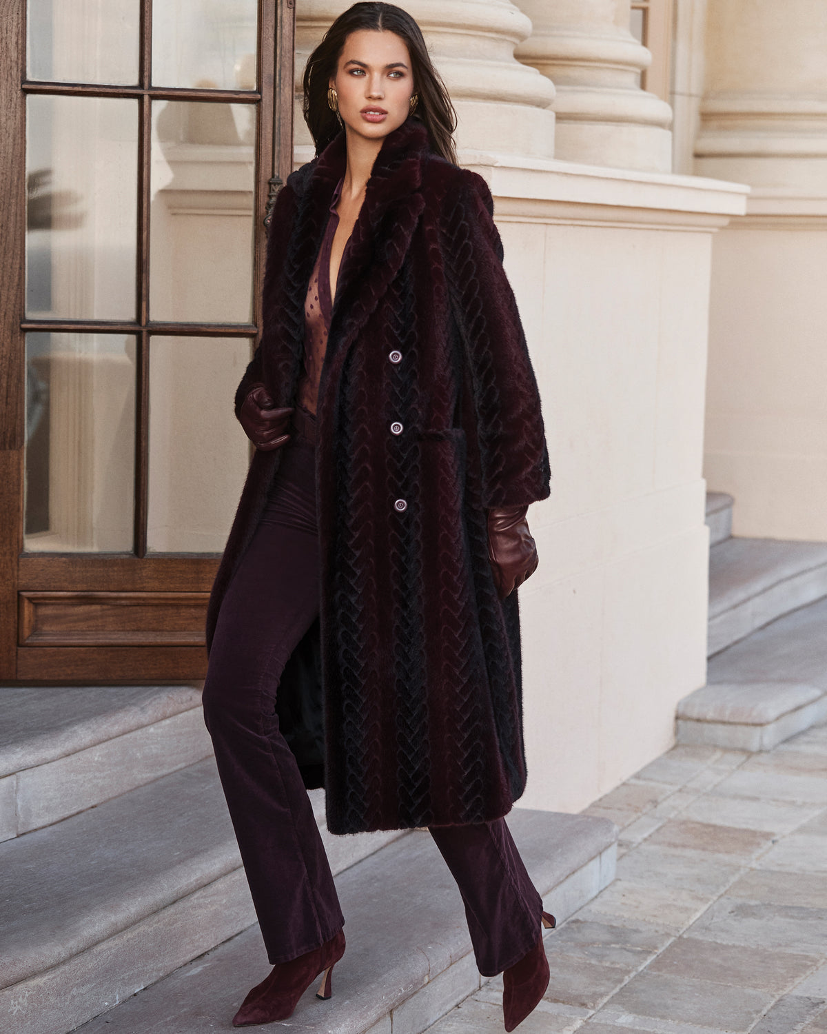 A woman in a dark burgundy patterned faux fur coat, matching trousers, and leather gloves stands on stone steps.