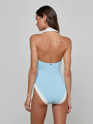A woman with long brown hair is seen from the back wearing the Natalya Collared One-Piece Swimsuit in light blue bouclé fabric with white trim and a halter neck, set against a plain gray background.
