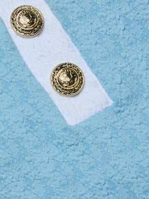Close-up of two ornate gold lion’s head buttons on the Natalya Collared One-Piece Swimsuit, shown on a white textured bouclé fabric against a light blue background.