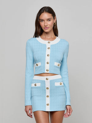 A woman wears the Adaline Cardigan Cover-Up, a powder blue and white textured two-piece with gold buttons, including a cropped jacket and mini skirt, standing against a plain light gray background.