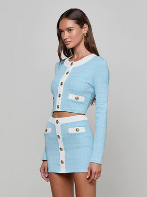 A woman stands against a plain gray background wearing the Adaline Cardigan Cover-Up—a powder blue, long-sleeve, cropped cardigan with white trim and gold buttons—paired with a matching mini skirt.