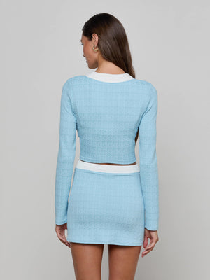 A woman with long brown hair stands with her back to the camera, wearing the Adaline Cardigan Cover-Up in powder blue. The cover-up features long sleeves and white trim, and she is set against a plain light gray background.