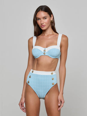 A woman wears the Alexandria Bikini Top in powder blue and white bouclé swim fabric with gold buttons. She stands against a light gray background, her long brown hair down and minimal makeup, looking toward the camera.