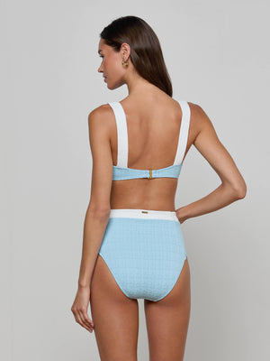 A woman with long brown hair poses against a plain background, wearing the Alexandria Bikini Top—a powder blue balconette style with thick white straps and a gold clasp—paired with matching high-waisted bottoms.