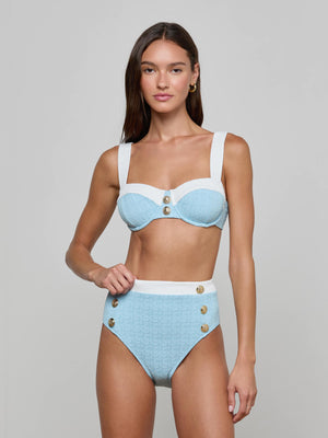 A woman stands against a plain background wearing the Vanessa High-Waisted Bikini Bottom in light blue and white bouclé swim fabric with gold buttons, offering moderate coverage and a supportive fit.