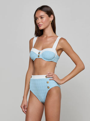 A woman stands against a plain background wearing the Vanessa High-Waisted Bikini Bottom, a light blue and white bikini with gold button details, wide straps, supportive fit, and crafted from bouclé swim fabric.