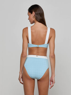 A woman with long brown hair is seen from the back wearing the Vanessa High-Waisted Bikini Bottom in light blue bouclé fabric, featuring wide white straps and waistband, offering moderate coverage against a plain light gray background.