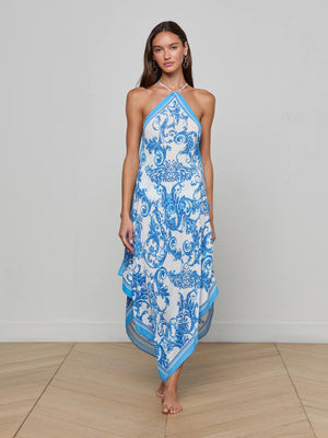 A woman stands barefoot on a wooden floor wearing the Elise Cover-Up Dress, a sleeveless halter midi with blue baroque print and an asymmetrical hem, set against a plain light gray background.