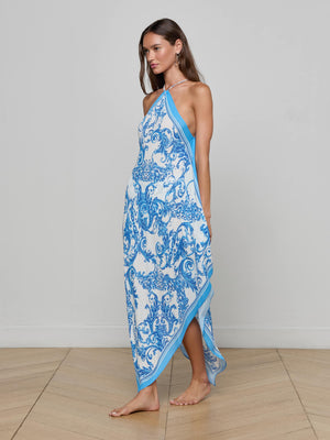 A woman stands barefoot on wooden flooring, wearing the Elise Cover-Up Dress—a blue baroque print halter midi made from water-safe crepe—set against a plain light gray wall.