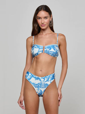A woman with long brown hair wears the Gianna Brazilian Bikini Bottom, a high-waisted, cheeky blue baroque print piece. She stands facing forward against a plain light gray background with a neutral expression.