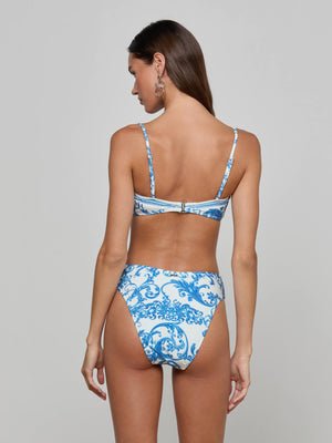 A woman with long brown hair models the Gianna Brazilian Bikini Bottom in a blue baroque print, featuring a cheeky cut and viewed from the back. She wears hoop earrings and stands against a plain light gray background.