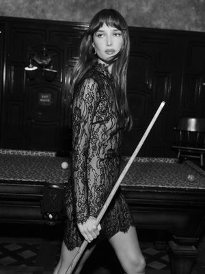 In a black and white photo, a woman wearing the Henrietta Lace Mini Dress with a high neckline stands by a pool table, holding a cue, in a dimly lit room with ornate wooden walls.