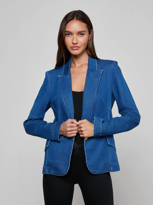 A woman with long brown hair faces forward, smiling slightly as she holds the lapels of the L’AGENCE Baldwin Blazer in stretch indigo denim, layered over a black top and pants against a plain light gray background.