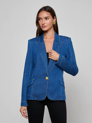 A woman with long brown hair models the L'AGENCE Baldwin Blazer, a blue stretch denim jacket with gold buttons, paired with black pants against a plain light gray background.