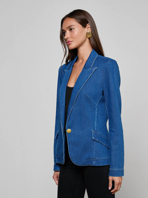 A woman with long brown hair models the L'AGENCE Baldwin Blazer in indigo wash with gold buttons, paired with a black top and black pants, standing against a plain light gray background.