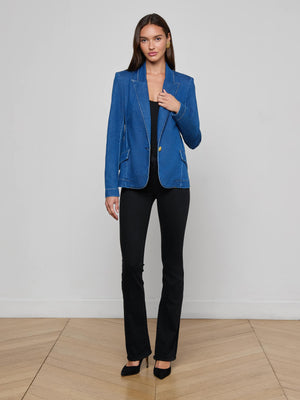 A woman stands indoors on a wooden floor, posing against a light gray wall while wearing the L'AGENCE Baldwin Blazer in an indigo wash, styled with a black top, black pants, and black heels.